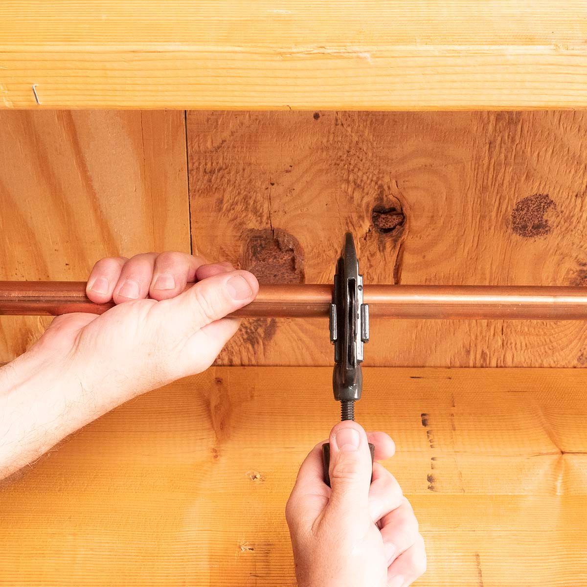 Hands grip a copper pipe while using a wrench to tighten a fitting against a wooden surface, indicating a plumbing task in a workshop.