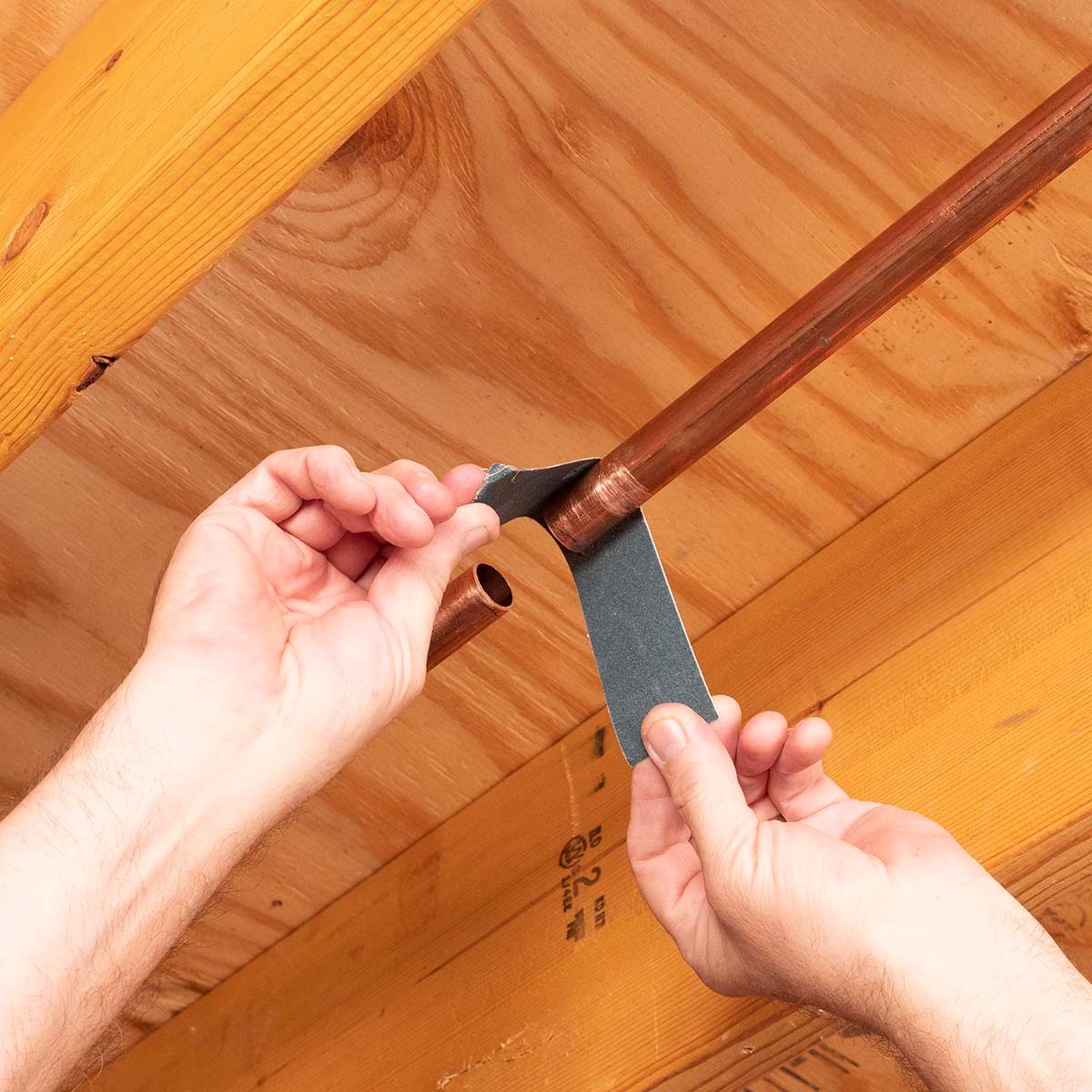 A person is sanding a copper pipe joint with sandpaper, positioned beneath wooden beams in a workshop or home setting.