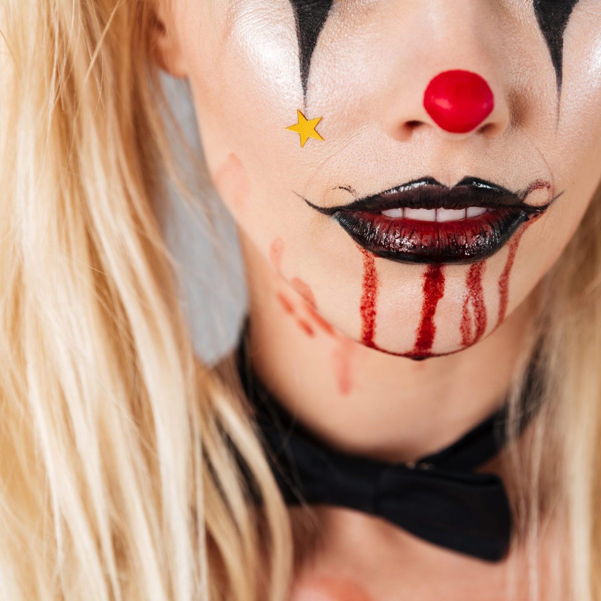 A clown's face is decorated with exaggerated makeup, featuring a red nose, black lips, and blood-like streaks, set against a light backdrop.