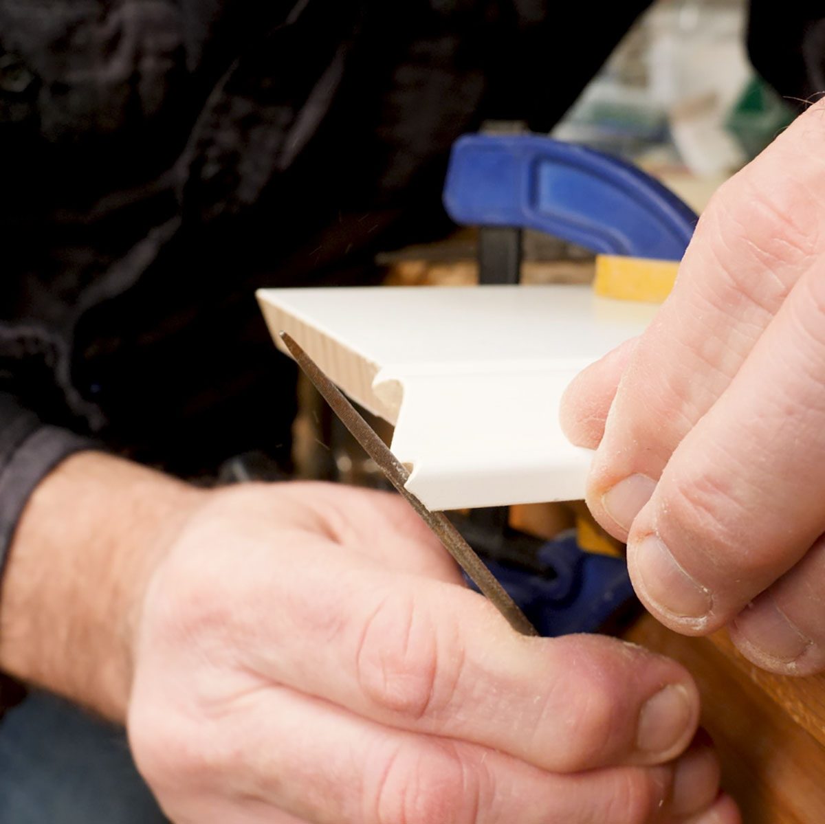 A hand holds a saw, cutting through a white, edge-shaped piece of wood secured by a blue clamp on a workbench.