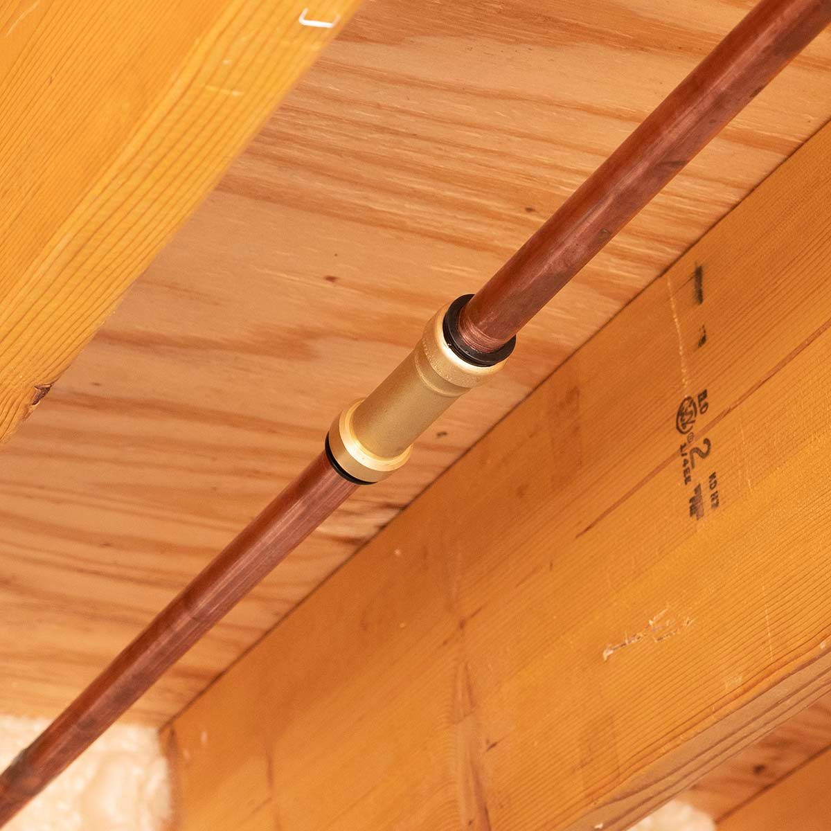 A copper pipe extends across a wooden ceiling, joined by a brass connector. The surrounding area features exposed wood beams and insulation material below.