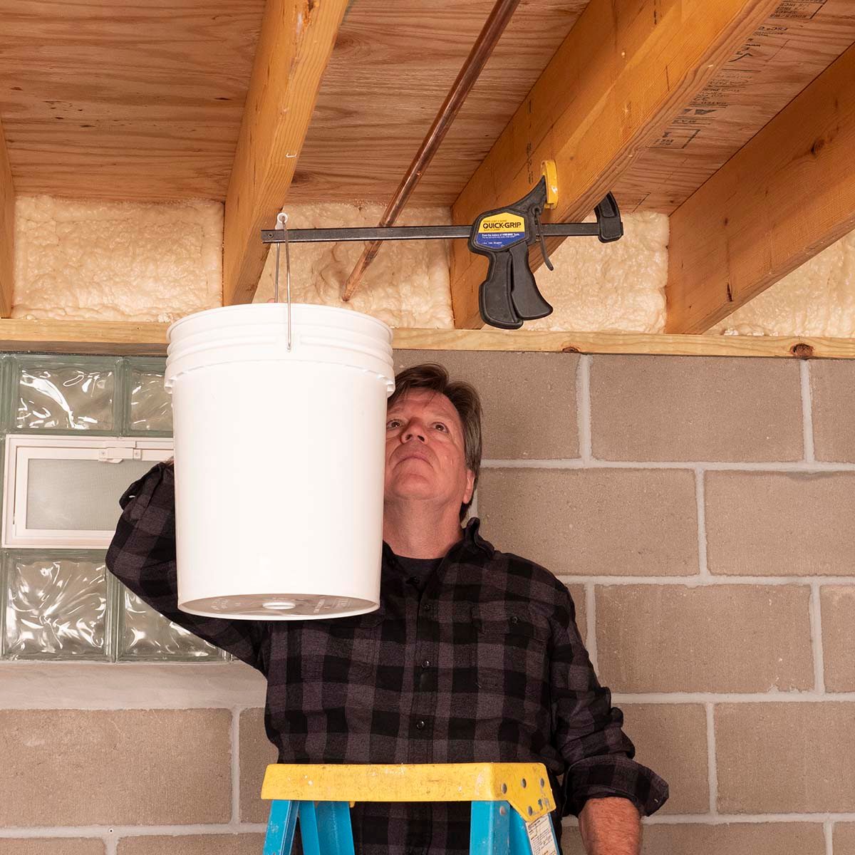 A man stands on a ladder, adjusting a white bucket suspended from the ceiling with a clamp, in a basement with concrete walls.