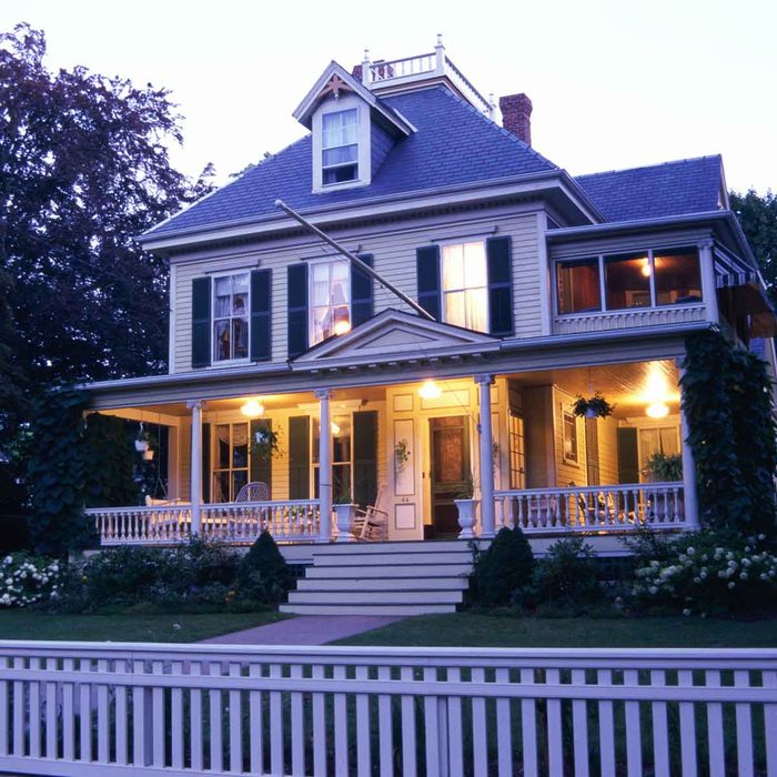 A large, well-lit Victorian house features a welcoming porch, surrounded by greenery and flowers, in a twilight setting.