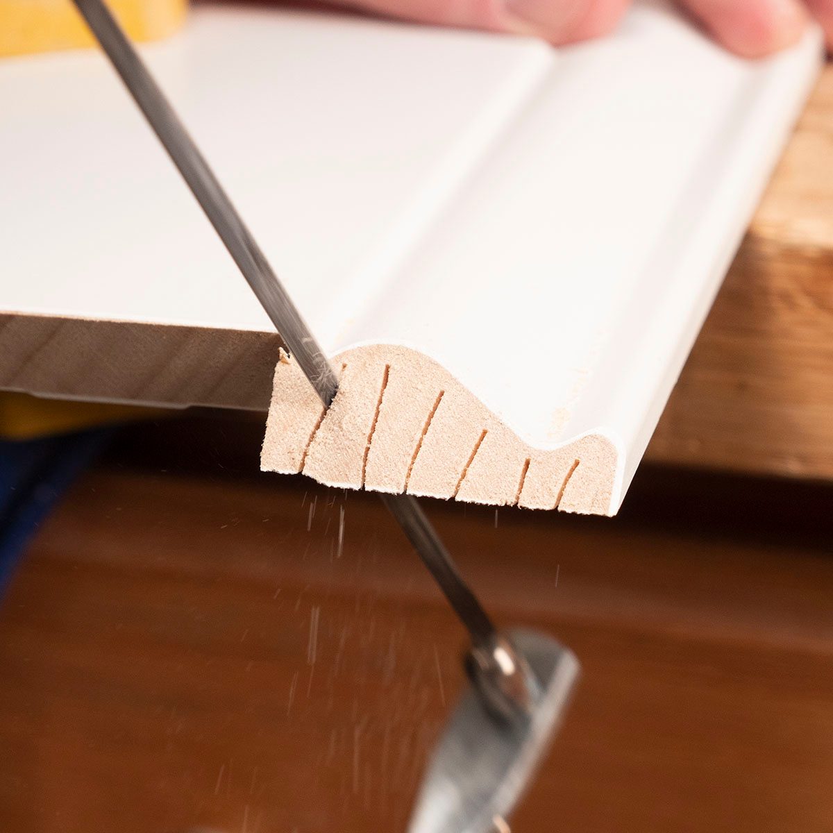 A saw cuts into the edge of a white molding piece, creating fine wood shavings, on a wooden workbench. A hand stabilizes the material.