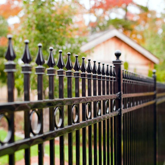 A black decorative fence stands upright, featuring pointed tips, enclosing a green garden area with trees and a wooden house in the background.