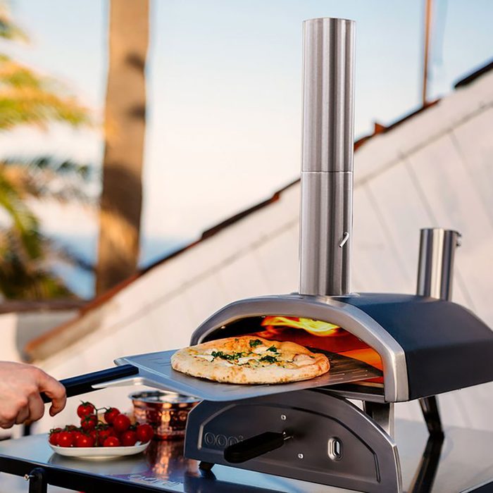 A pizza is being removed from a sleek outdoor pizza oven, surrounded by a sunny patio with palm trees and fresh tomatoes on a nearby plate.