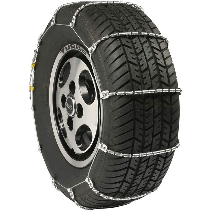 A black tire with a patterned tread is fitted with metallic chains, suggesting preparation for icy conditions, possibly on a vehicle.