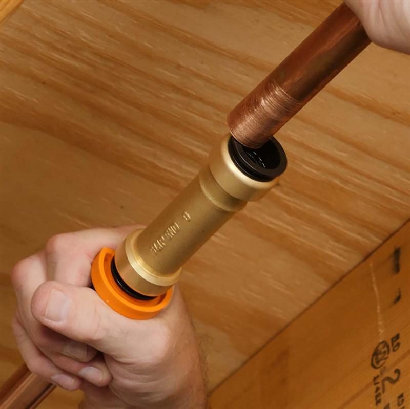 A hand connects a copper pipe to a fitting with an orange ring, within a wooden environment, indicating a plumbing or installation task.