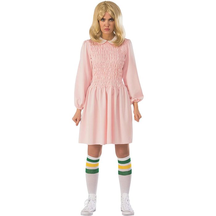 A person stands confidently in a pink, smocked dress, white sneakers, and knee-high socks with green and yellow stripes, set against a plain white background.
