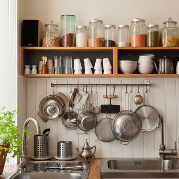 Shelves display jars of food. Hanging kitchen utensils are organized near a sink. A potted plant adds greenery to the cozy kitchen setting.