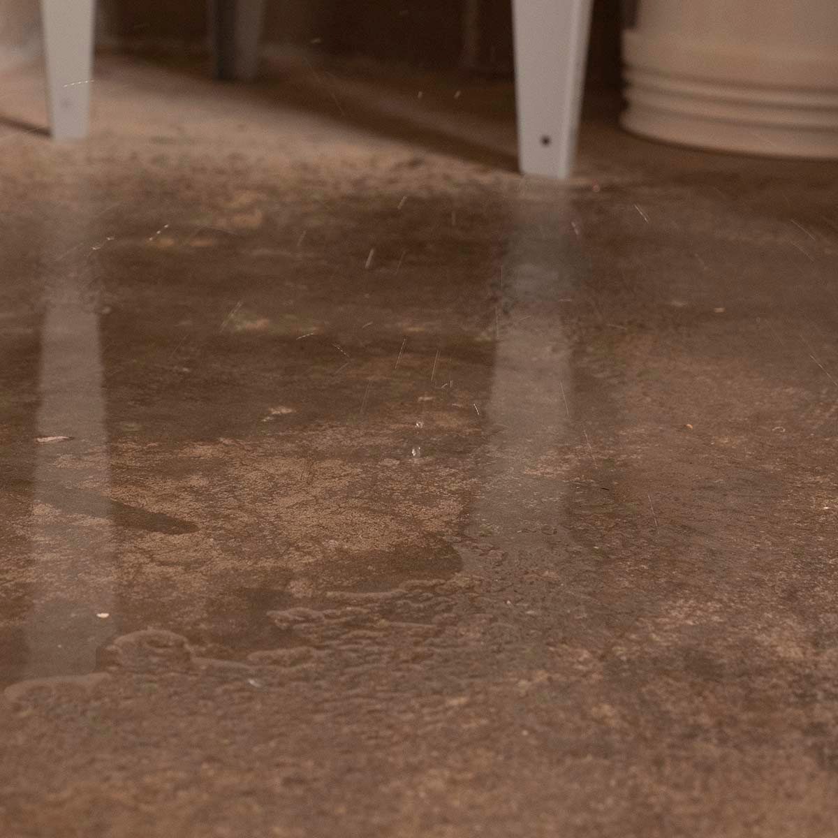 Water drips onto a stained, concrete floor, creating small splashes, while faint reflections of nearby objects are visible in the water's surface.