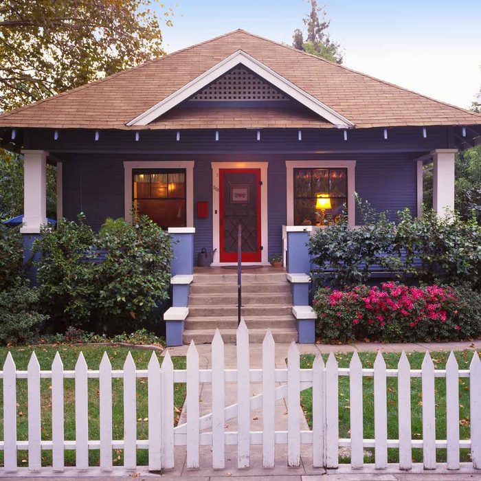 A blue house with a red door stands surrounded by green bushes and pink flowers, featuring white picket fencing and a welcoming entrance with steps.