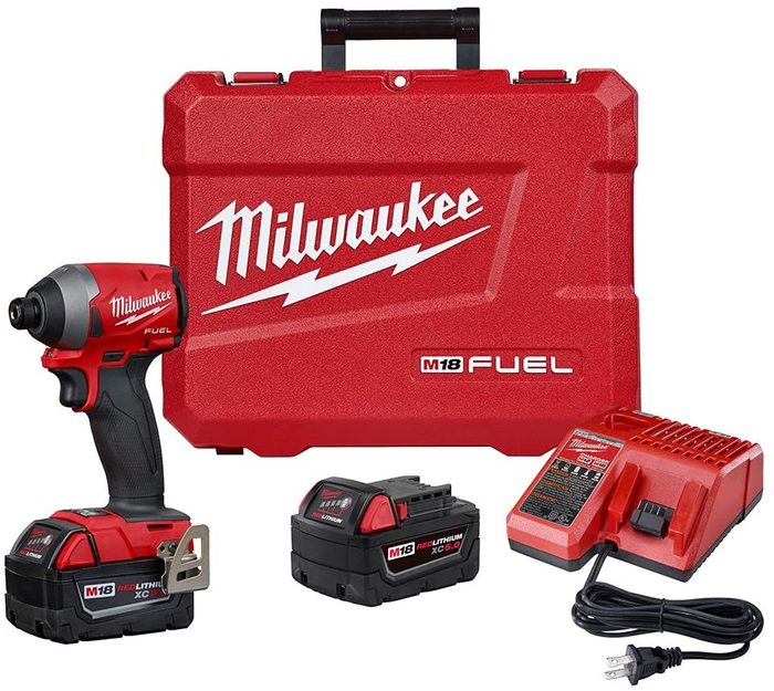 A red Milwaukee impact driver is positioned next to its charging station and two batteries, all packaged in a matching red carrying case.
