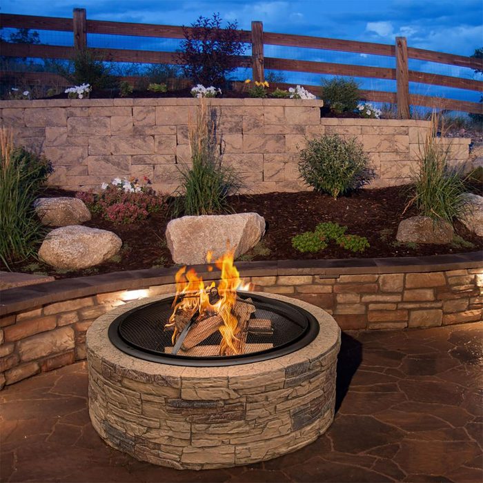 A stone fire pit burns logs, creating flames. It's surrounded by a landscaped yard with plants and rocks, under a twilight sky with a wooden fence.