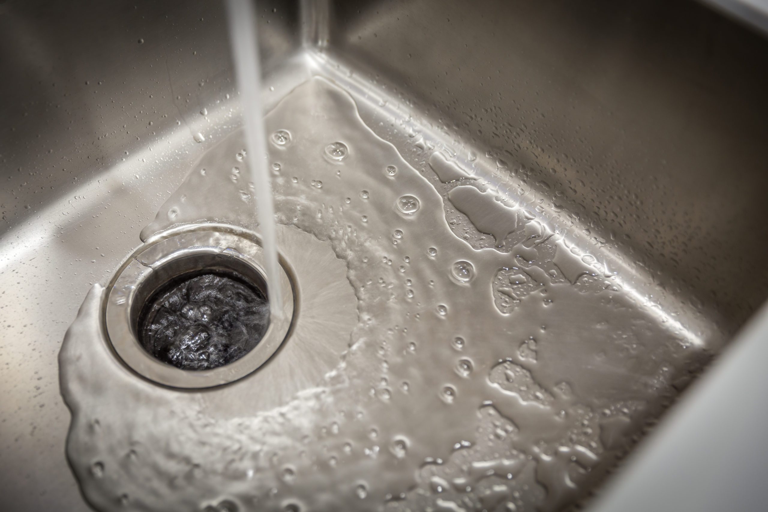 A stream of water flows into a stainless steel sink, creating bubbles around a circular drain at the bottom, with droplets on the sink's surface.