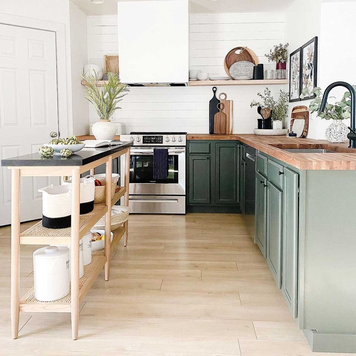 A kitchen features a dark countertop island displaying plants, while green cabinets and stainless steel appliances create a modern, clean space with wooden decor.