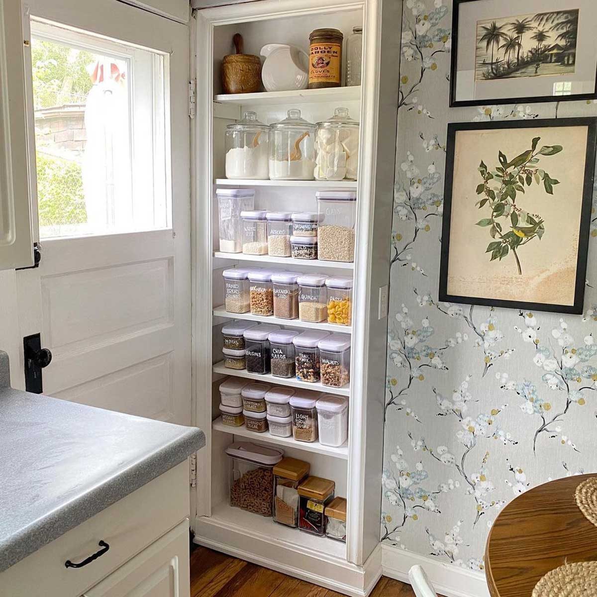 A neatly organized pantry contains various transparent containers of grains and spices. The background features floral wallpaper and a wooden table with a light surface.