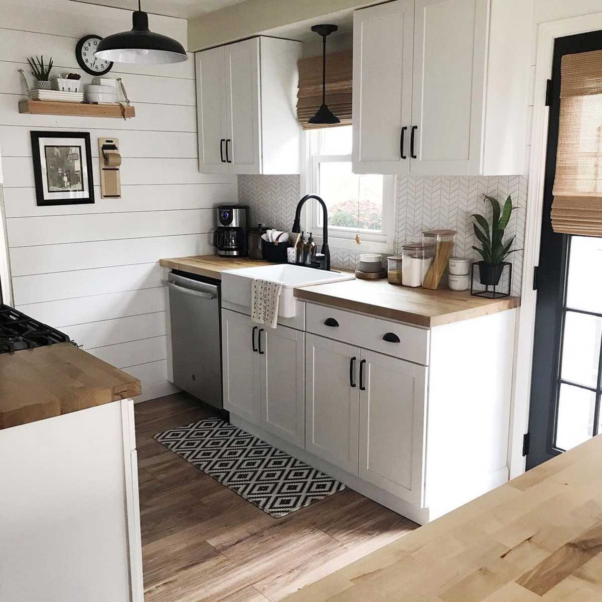 A modern kitchen features white cabinets and a wooden countertop, with a coffee maker on display. Natural light enters through a window beside a potted plant.