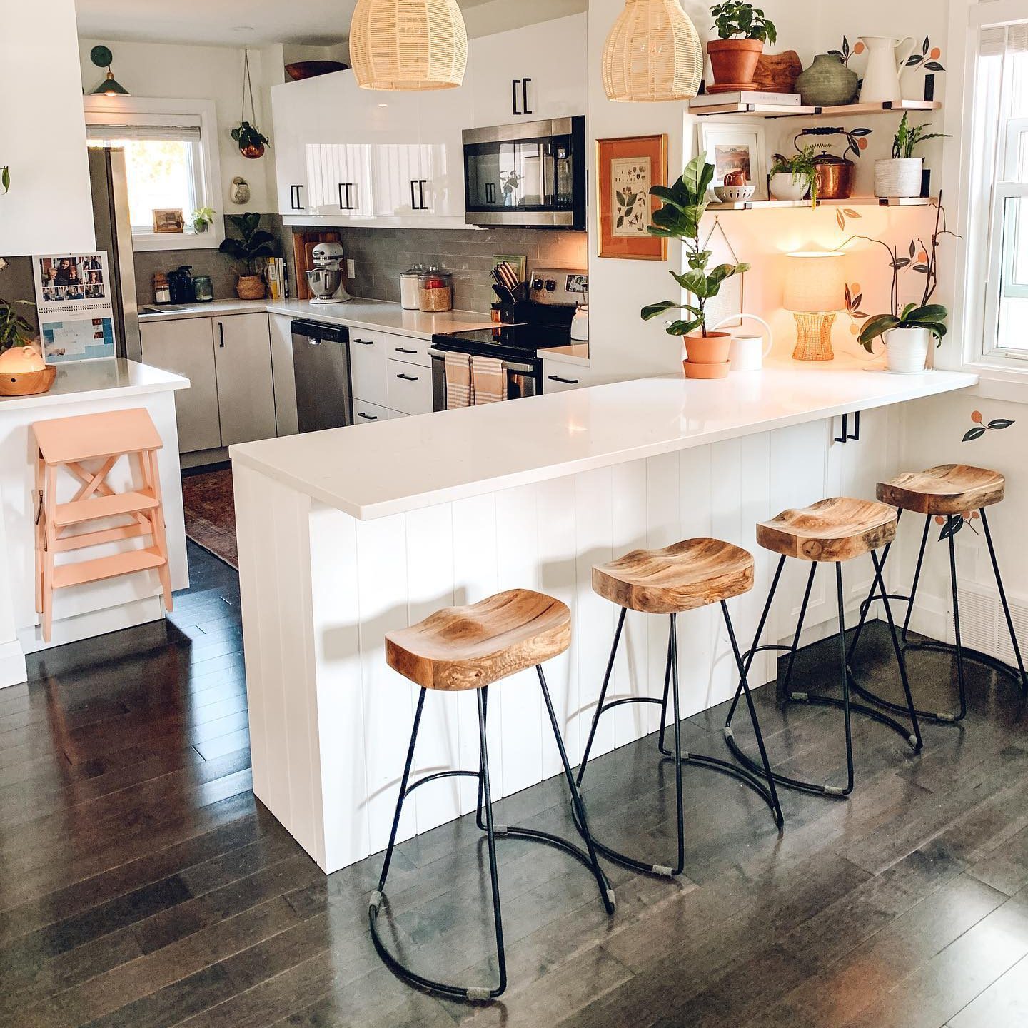 A modern kitchen features a white countertop with four wooden stools, while light streams through a window, enhancing the welcoming atmosphere.