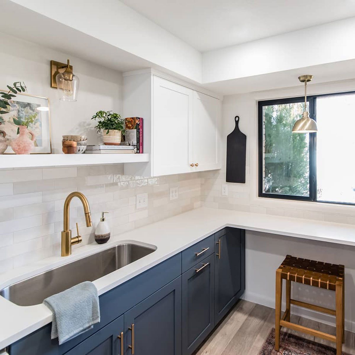 A kitchen features a stainless steel sink and gold faucet, with white cabinets and shelves displaying plants and decor, illuminated by a pendant light above.