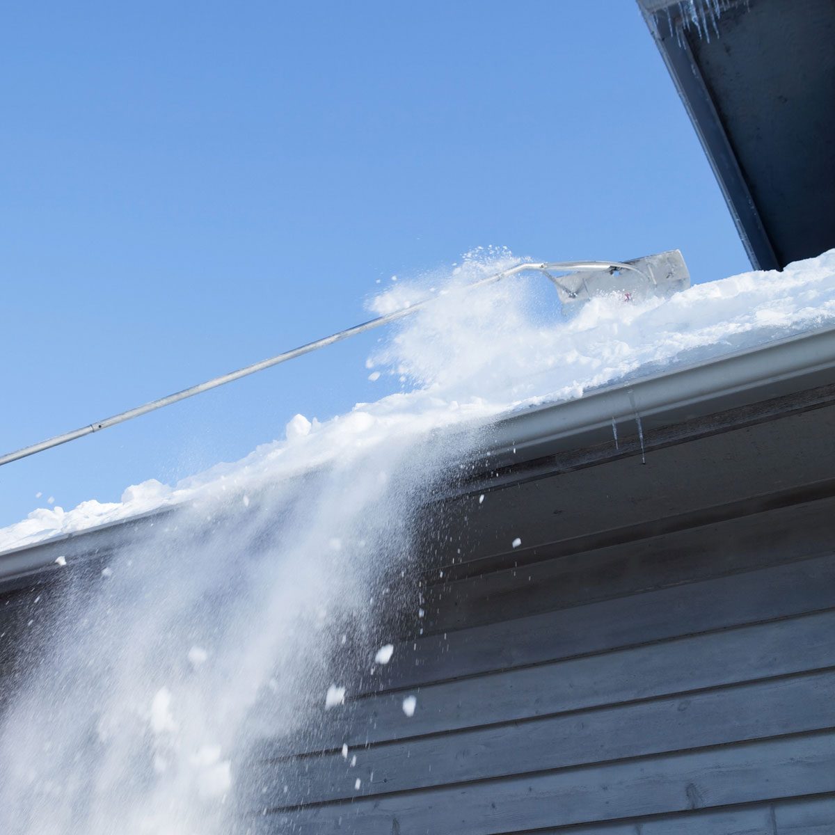 A snow shovel removes snow from a roof, with flakes cascading downwards against a bright blue sky and a wooden building below.