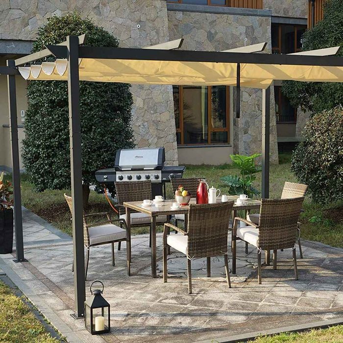 A patio set with a table and chairs is surrounded by greenery, featuring a grill nearby, under a shaded pergola in a residential setting.
