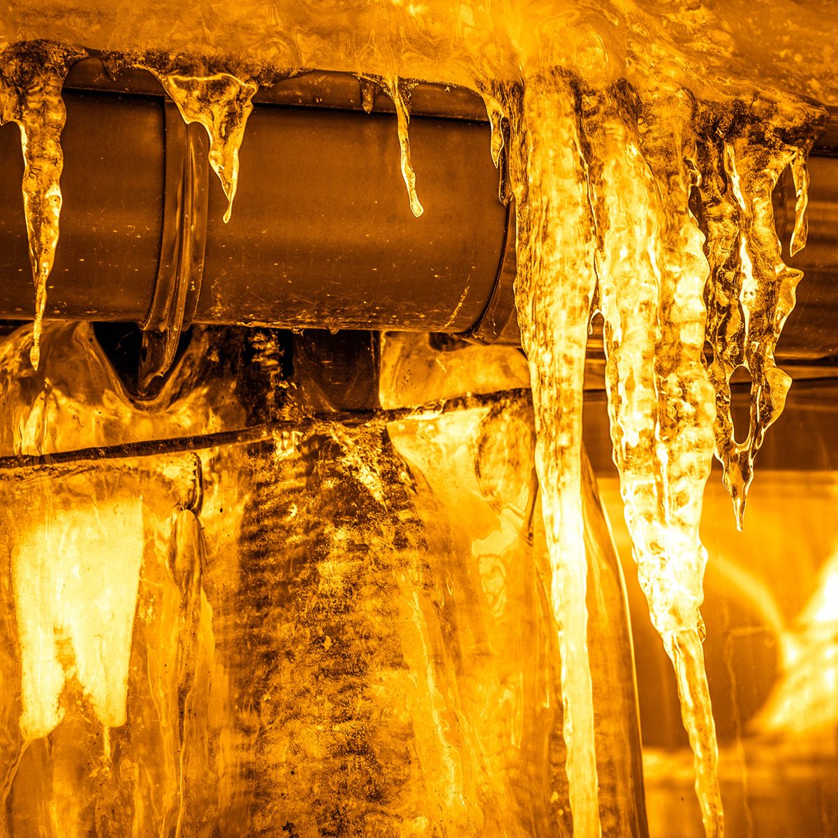 Icicles hang from a dark metal pipe, dripping water in a warm, amber-lit environment, creating a striking contrast between the cold ice and warm colors.