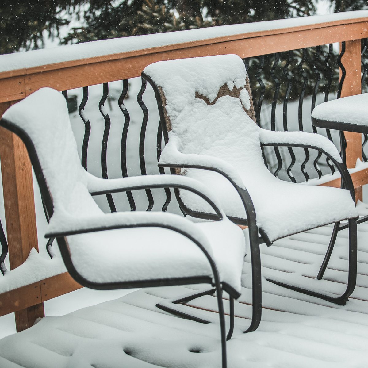 Two snow-covered chairs sit on a snow-blanketed deck, surrounded by a wooden railing against a backdrop of falling snow.