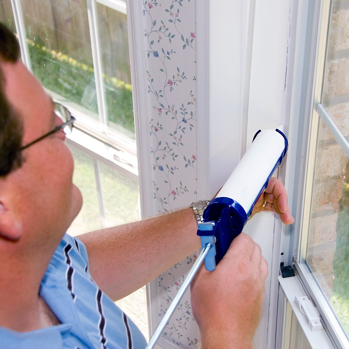 A man uses a caulking gun to seal a window frame. The interior features floral wallpaper and sunlight illuminates the scene through the window.