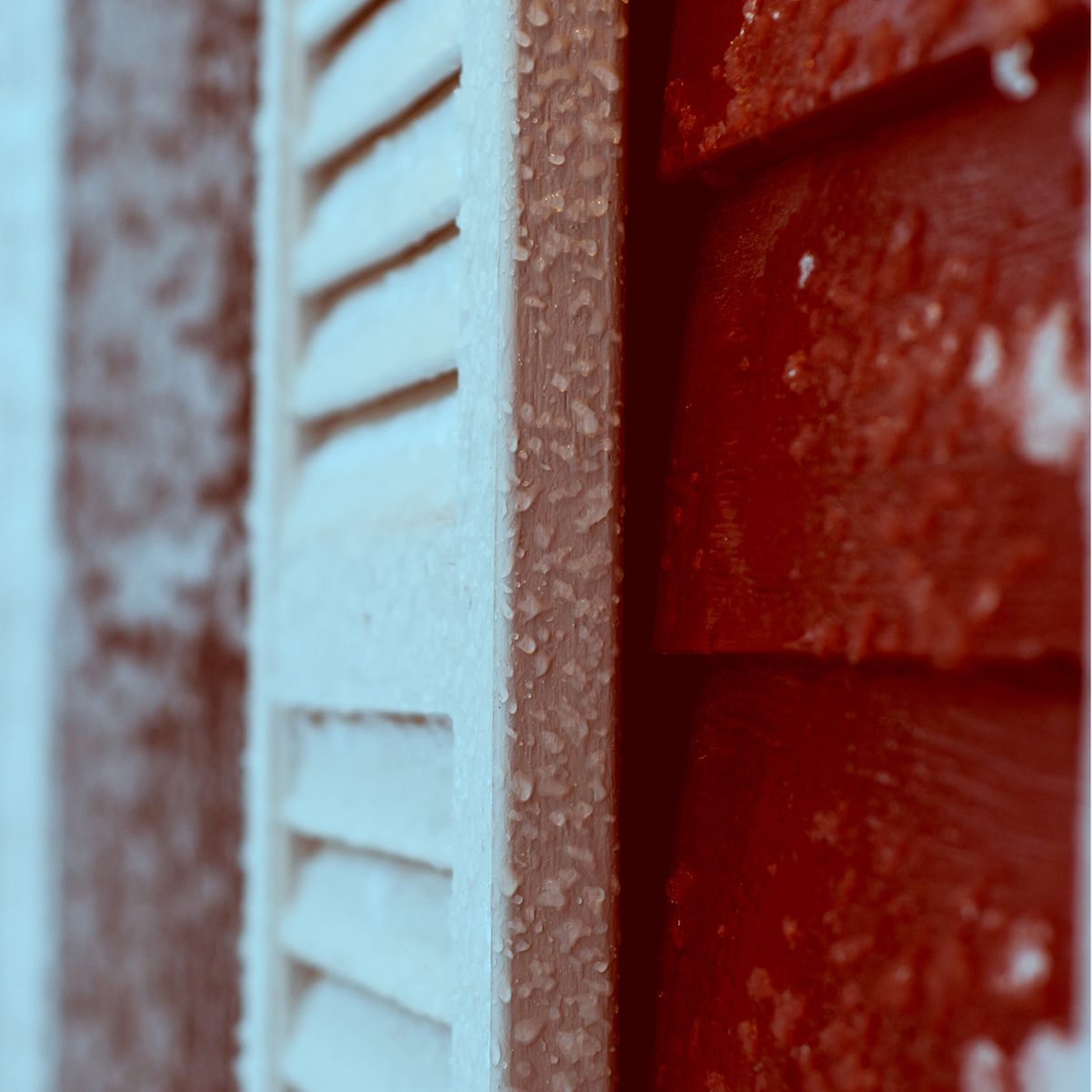 A white shutter and a red wall are coated in frost, with icicles forming on the surfaces, suggesting a cold, winter environment.