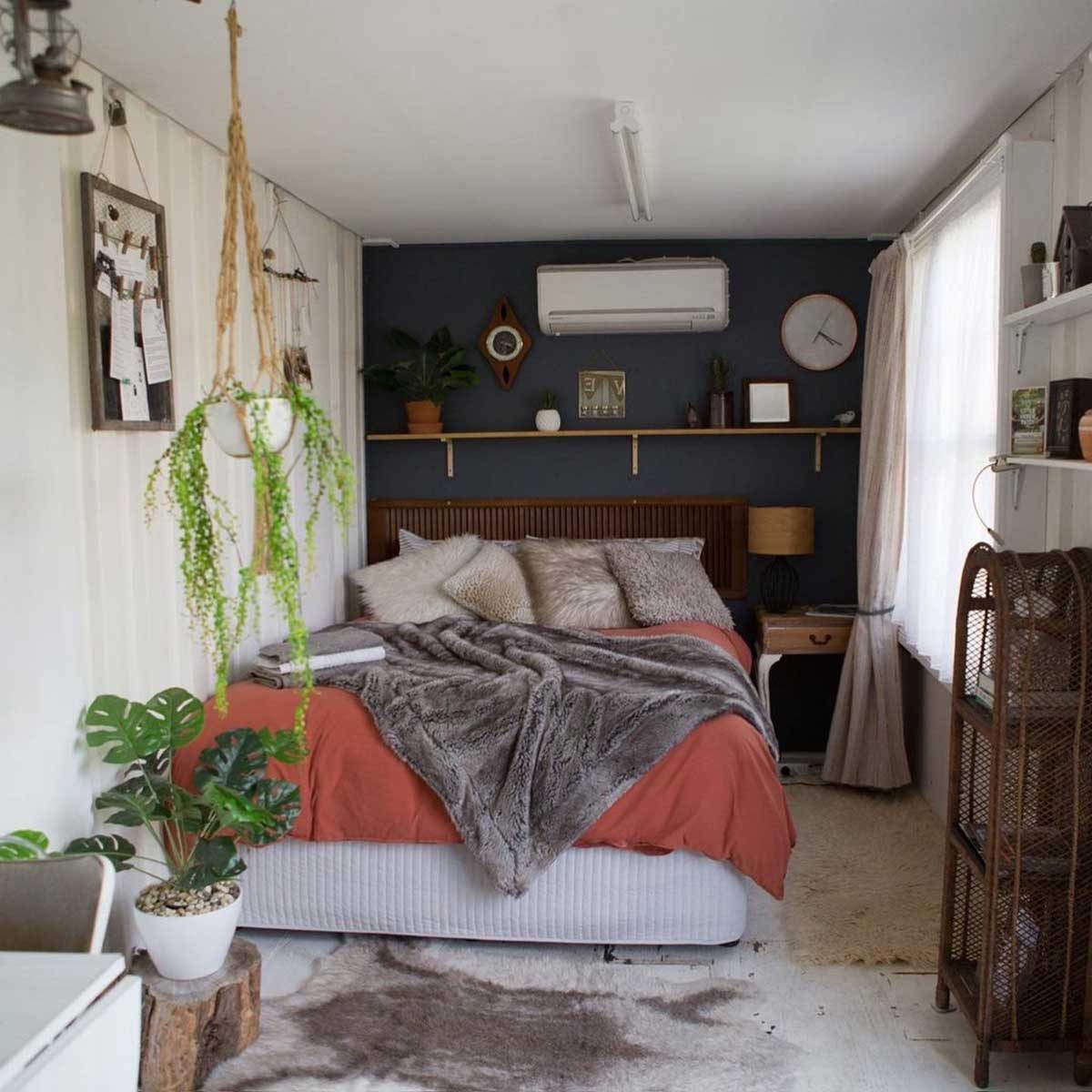 A cozy bedroom features a bed with soft linens and decorative pillows, surrounded by plants, shelves, and soft lighting, creating a warm, inviting atmosphere.