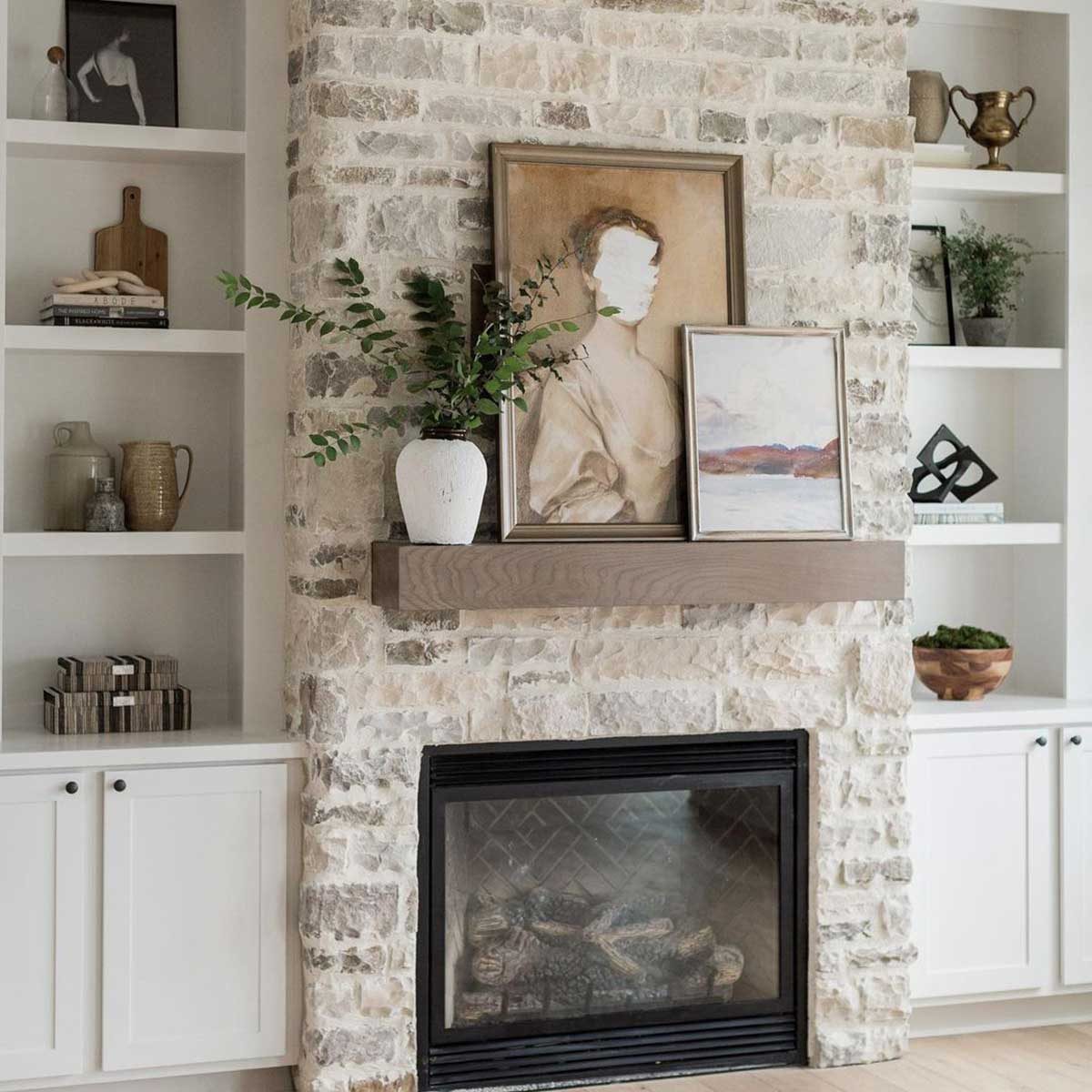 A mantel displays two frames and a vase with foliage above a stone fireplace, complemented by shelves holding various decorative items in a cozy room.