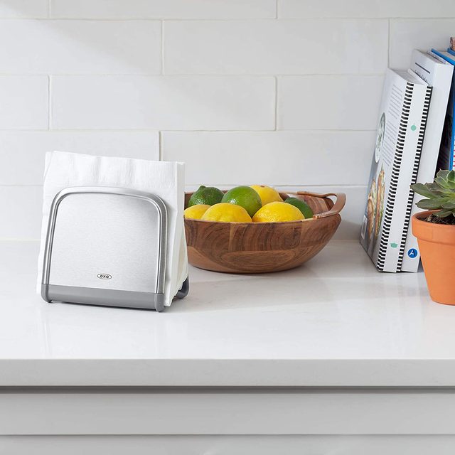 A napkin holder stands on a countertop, holding white napkins. Nearby, a wooden bowl contains lemons and limes, with books and a potted plant in the background.
