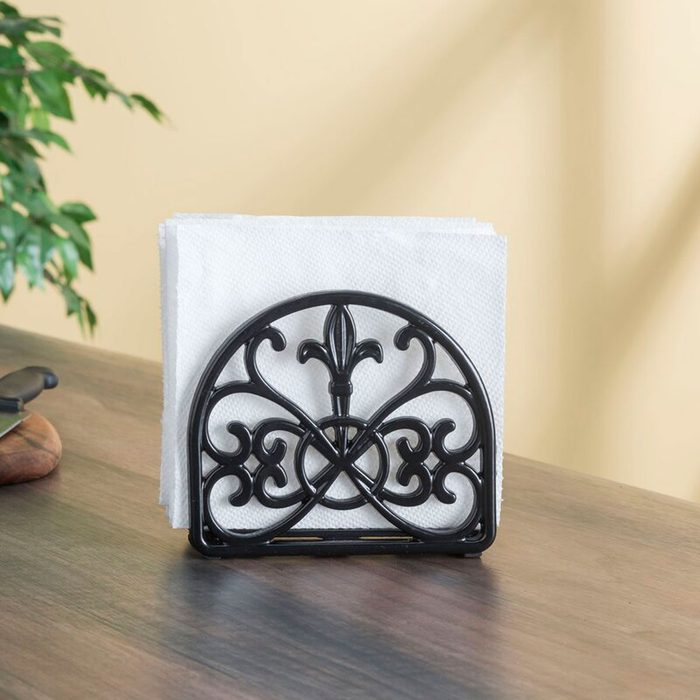 A decorative black napkin holder supports white napkins on a wooden table, set against a light yellow wall with a green plant nearby.