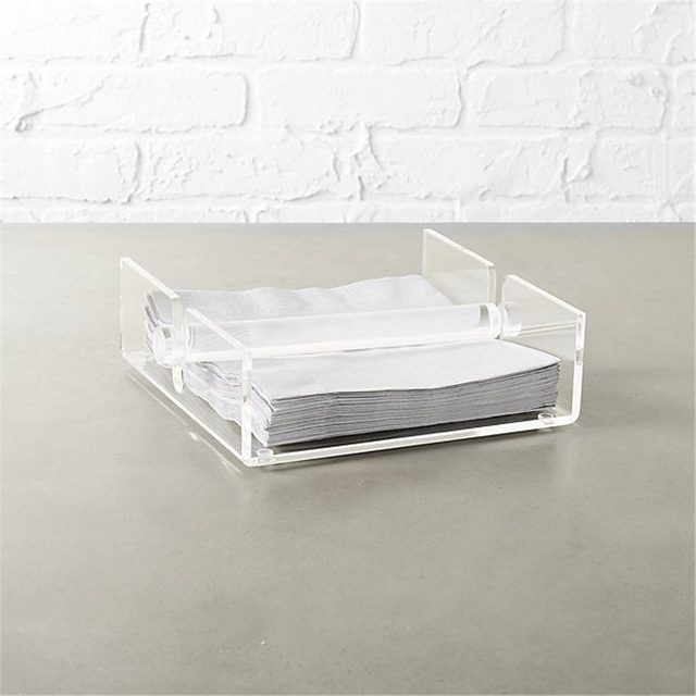 A stack of gray napkins is neatly stored in a clear acrylic holder on a smooth, light-colored surface, with a textured white brick wall in the background.