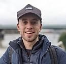 A young man smiles, wearing a cap and jacket, standing outdoors with a cloudy sky and a body of water in the background.