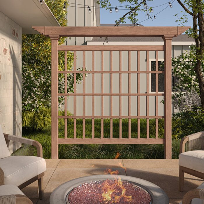 A wooden trellis stands in a backyard, surrounded by greenery. Nearby, a fire pit with flames flickers on a stone patio, creating a cozy atmosphere.