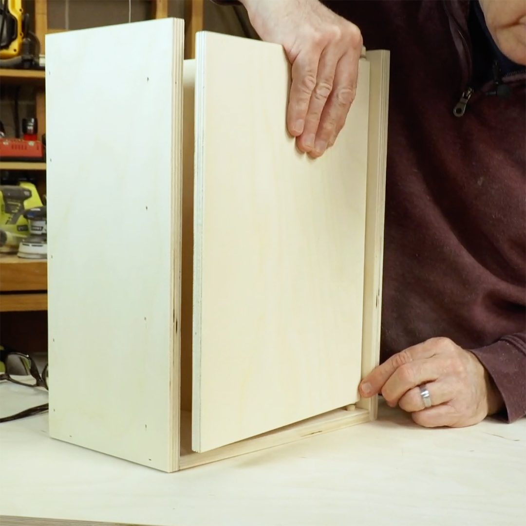 A hand places a wooden panel into a structure while assembling a cabinet in a workshop, surrounded by tools and materials on a workbench.