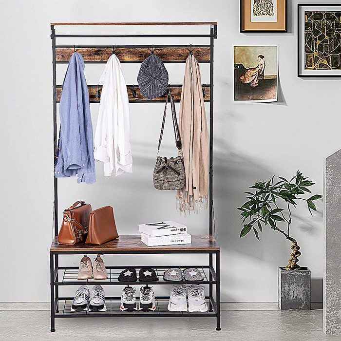 A coat rack holds various clothing items and bags, while shoes and boxes are arranged on a storage bench against a light wall, near a plant.