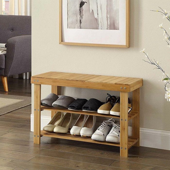 A wooden shoe rack displays various pairs of shoes and slippers, positioned against a beige wall in a well-lit room with a framed picture above it.