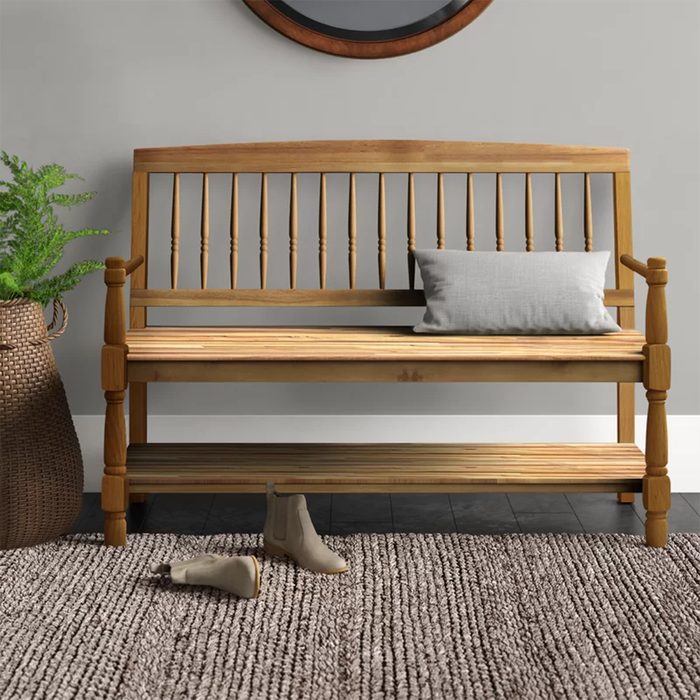 A wooden bench sits against a gray wall, displaying a gray pillow, while casual boots lie on a textured rug nearby. A potted plant adds greenery.