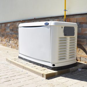 A large white generator sits on a concrete base beside a stone wall, with a yellow pipe connected above, in bright sunlight.