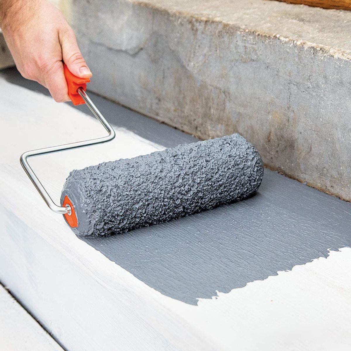 A hand rolls a textured gray paint roller over a freshly painted surface, with a concrete background and partially painted white areas nearby.