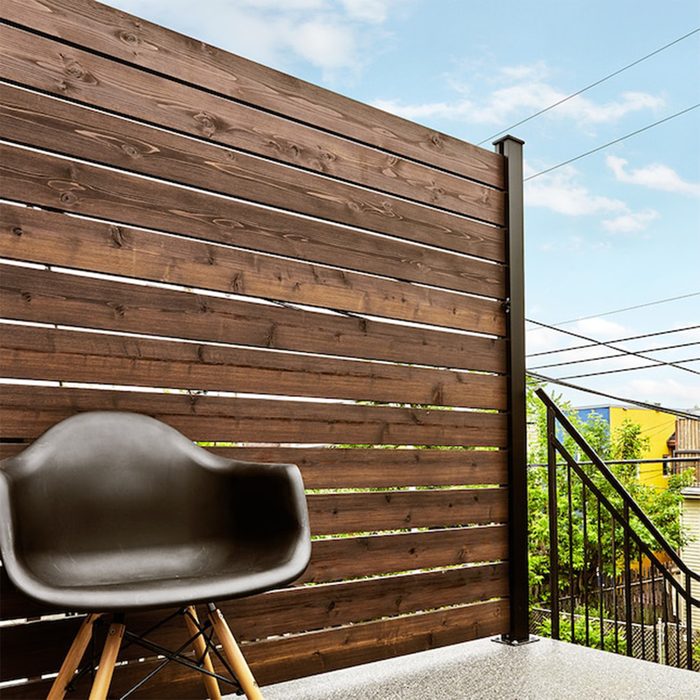 A black plastic chair sits on a balcony, adjacent to a wooden slatted wall, overlooking a neighborhood with greenery and power lines against a blue sky.