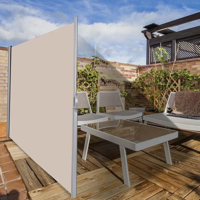 An outdoor seating area features a beige privacy screen beside white chairs and a glass table, surrounded by plants and wooden decking under a blue sky.