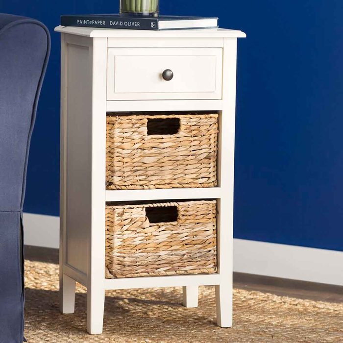 A white nightstand with a drawer and two woven baskets stands on a textured rug against a blue wall, topped with a book and a plant.