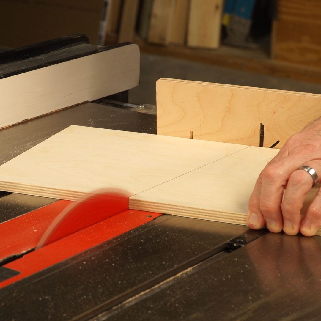 A hand guides a wooden board through a table saw, where the blade cuts it along a marked line in a workshop environment.