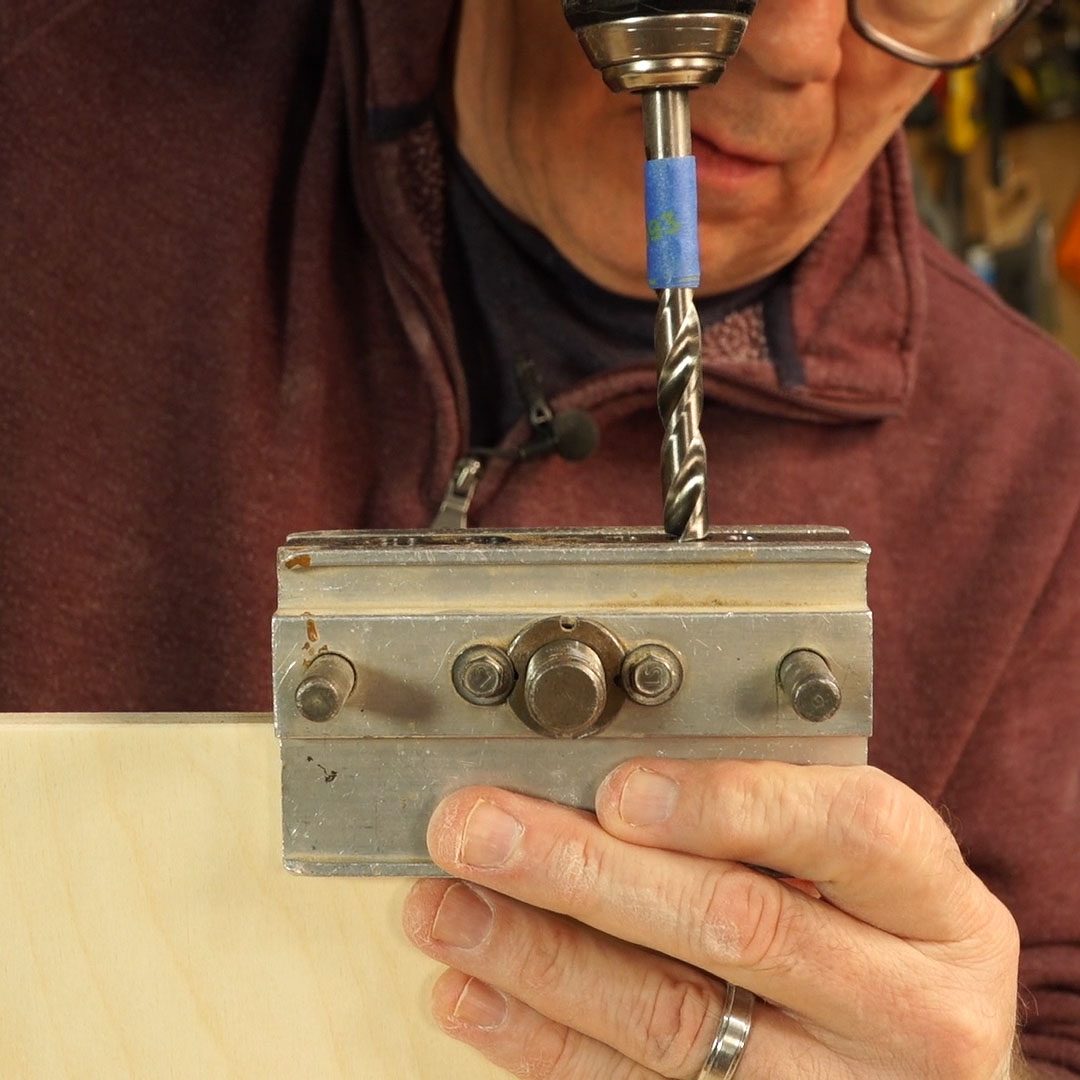 A person uses a drill on a metallic guide, positioning it over a wooden surface to create precise holes in a workshop environment.