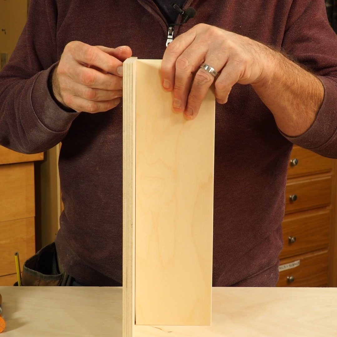 Hands are positioning a wooden panel upright on a work surface, surrounded by tools and woodcraft materials in a workshop environment.