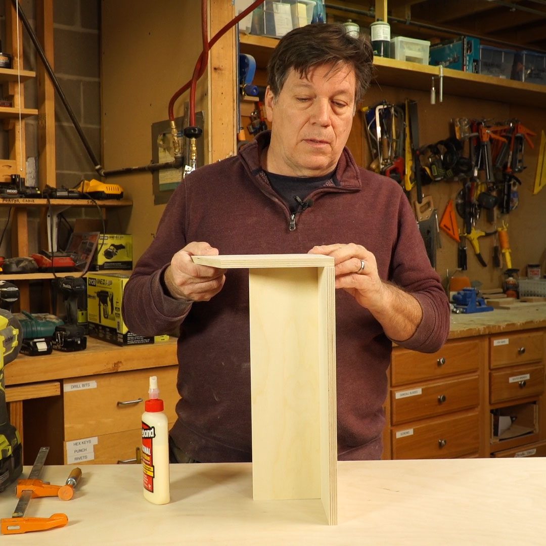 A person holds a vertical wooden piece, applying glue in a workshop filled with tools and equipment, focused on assembling a project.